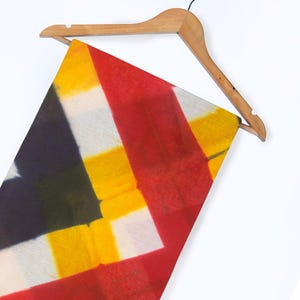 Puede incluir: Una colorida bufanda con un patrón geométrico de rayas rojas, amarillas, blancas y negras, colgada de una percha de madera. El diseño de la bufanda es una composición abstracta audaz.