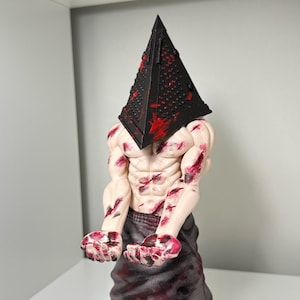 Könnte beinhalten: Eine 3D-gedruckte Figur eines Charakters aus der Silent Hill-Videospielserie. Der Charakter trägt einen schwarzen, pyramidenförmigen Kopf mit roten Akzenten und eine graue, blutige Robe. Die Figur steht auf einem schwarzen Sockel mit den Worten "Silent Hill" und "3D GadgeTry" darauf.