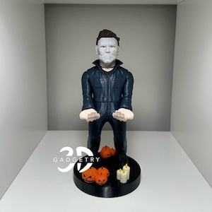 May include: A 3D printed figurine of Michael Myers from the Halloween franchise. The figurine is wearing a blue jumpsuit and a white mask. The figurine is standing on a black base with small pumpkins and candles.