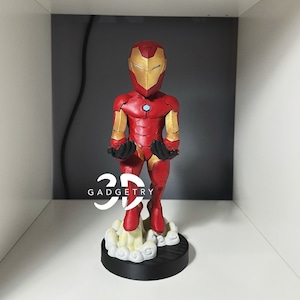 May include: A red and gold Iron Man figurine with a white cloud base. The figurine is standing with its arms outstretched. The text "GADGETRY" is visible on the base.