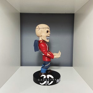 K&ouml;nnte beinhalten: Eine rot-blaue Bobblehead-Figur eines Charakters mit einem sch&auml;delartigen Kopf und einem Rucksack. Die Figur steht auf einem schwarzen Sockel mit dem aufgedruckten Text "3D GADGETRY IRON".