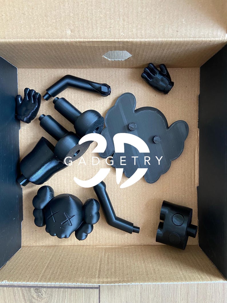 May include: A black 3D printed figure of a cartoon character with a skull and crossbones on its head. The figure is disassembled and comes in multiple pieces, including the head, arms, legs, and a base. The pieces are in a cardboard box.