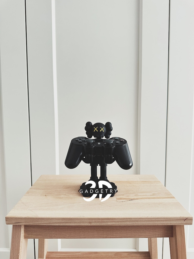 May include: Black and yellow 3D printed phone stand shaped like a video game controller with a black and yellow figure on top. The stand is on a wooden table and has the text "3D GADGETRY" on the base.