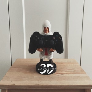 May include: A black PlayStation 3 controller is held by a white figurine of a hooded figure. The figurine is standing on a black base with the text "3D GADGETRY" printed on it.