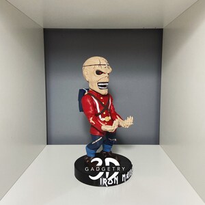 K&ouml;nnte beinhalten: Eine rote und schwarze 3D-Wackelfigur eines Charakters mit einem Sch&auml;delgesicht und einer rot-blauen Uniform. Die Wackelfigur steht auf einem schwarzen Sockel mit dem Text "3D Gadgetry Iron Maiden" in wei&szlig;.