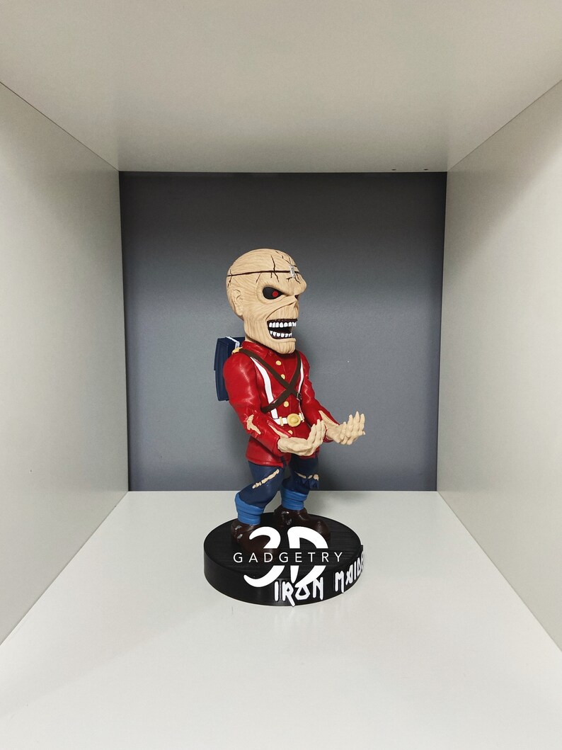 K&ouml;nnte beinhalten: Eine rote und schwarze 3D-Wackelfigur eines Charakters mit einem Sch&auml;delgesicht und einer rot-blauen Uniform. Die Wackelfigur steht auf einem schwarzen Sockel mit dem Text "3D Gadgetry Iron Maiden" in wei&szlig;.
