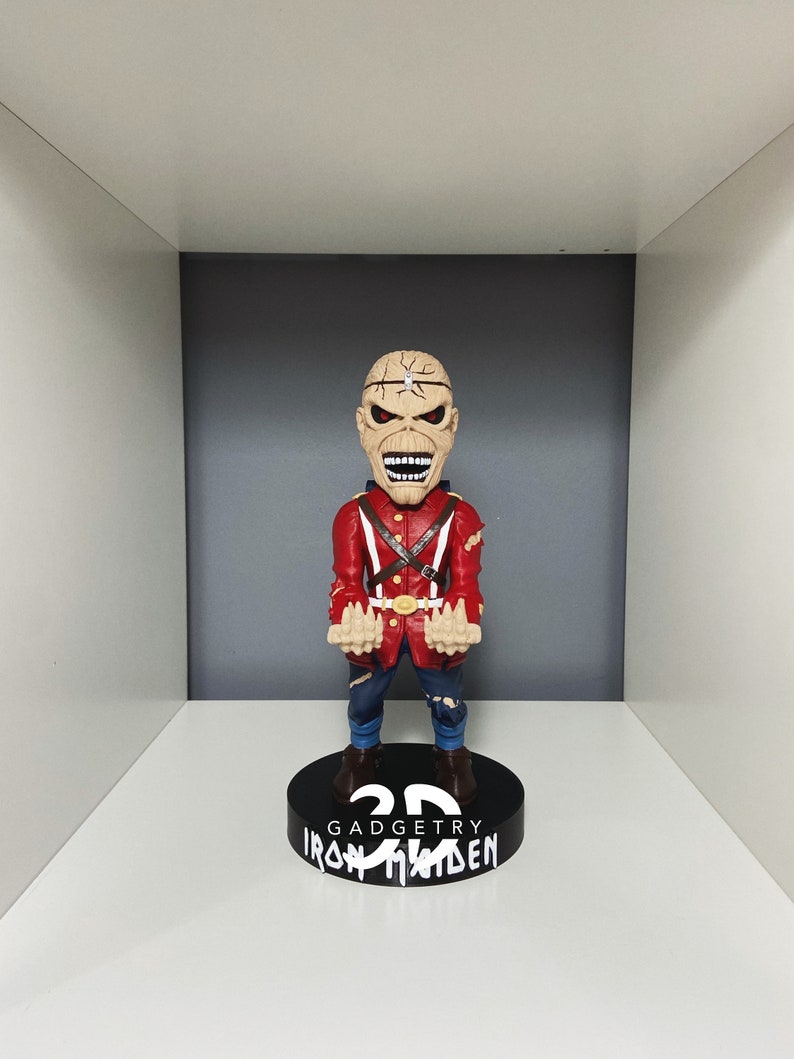 K&ouml;nnte beinhalten: Eine rot-schwarze Bobblehead-Figur eines Mannes mit einem Metallgesicht und einer rot-wei&szlig;en Uniform. Die Figur steht auf einem schwarzen Sockel mit der Aufschrift "3D GadgeTry Iron Maiden".