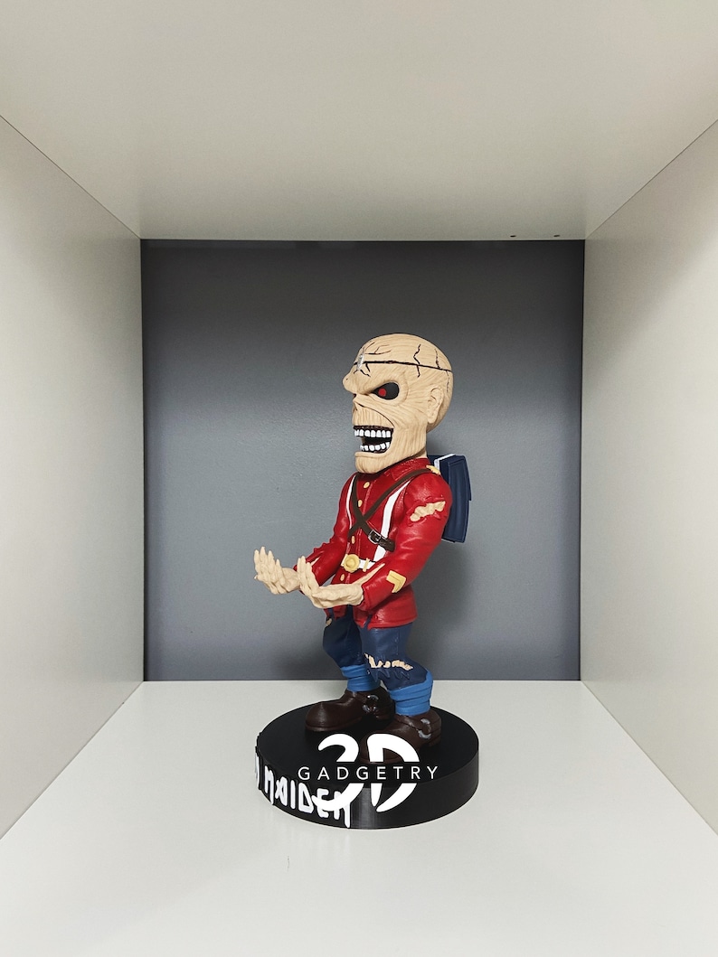 K&ouml;nnte beinhalten: Eine rot-blaue 3D-gedruckte Figur eines Charakters mit einem sch&auml;delartigen Gesicht. Die Figur steht auf einem schwarzen Sockel mit dem aufgedruckten Text "3D GADGETRY" und "MAIDEN".
