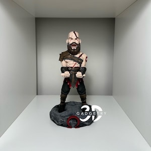 Puede incluir: Una figura Funko Pop de Kratos con un atuendo rojo y negro, barba y un hacha roja y negra. La figura está de pie sobre una base de roca gris con la letra griega Omega.