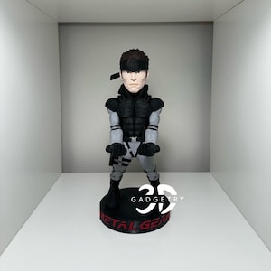 May include: A black and grey figurine of a character from the Metal Gear video game series. The figurine is posed in a fighting stance with a black headband and a grey and black outfit. The base of the figurine is black with the words "Metal Gear" in red.
