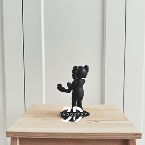May include: Black figurine of a cartoon character with two buns on top of its head. The figurine is standing on a wooden table with the text "3D GADGETRY" printed on the surface.