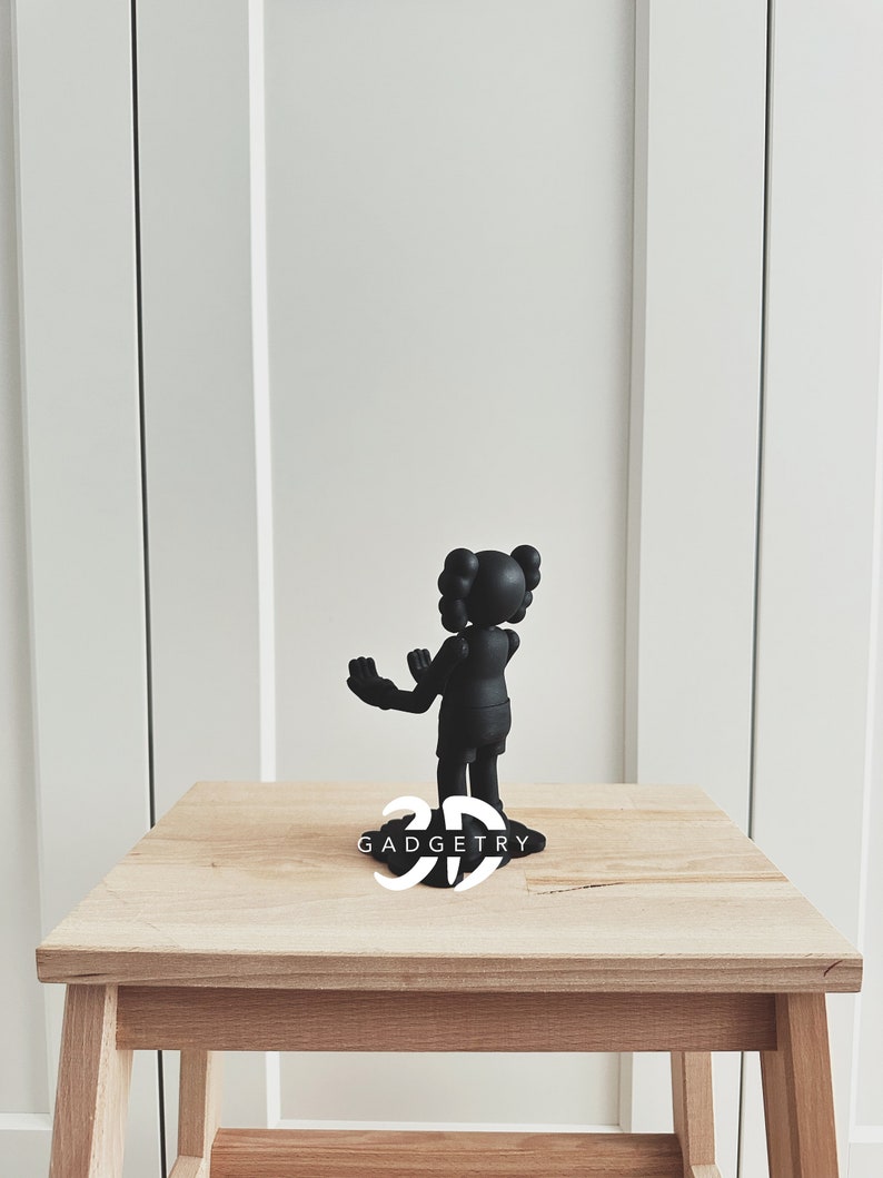 May include: Black figurine of a cartoon character with two buns on top of its head. The figurine is standing on a wooden table with the text "3D GADGETRY" printed on the surface.