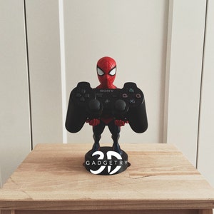 Può includere: Una statuetta di Spider-Man rossa e blu che tiene un controller nero Sony PlayStation 3. La statuetta è in piedi su una base nera con il testo "3D GADGETRY" stampato su di essa.