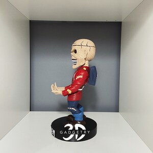 K&ouml;nnte beinhalten: Eine rot-wei&szlig;e Bobblehead-Figur eines Skeletts, das eine rote Jacke, blaue Jeans und einen blauen Rucksack tr&auml;gt. Die Figur steht auf einem schwarzen Sockel mit dem Text "3D GADGETRY".