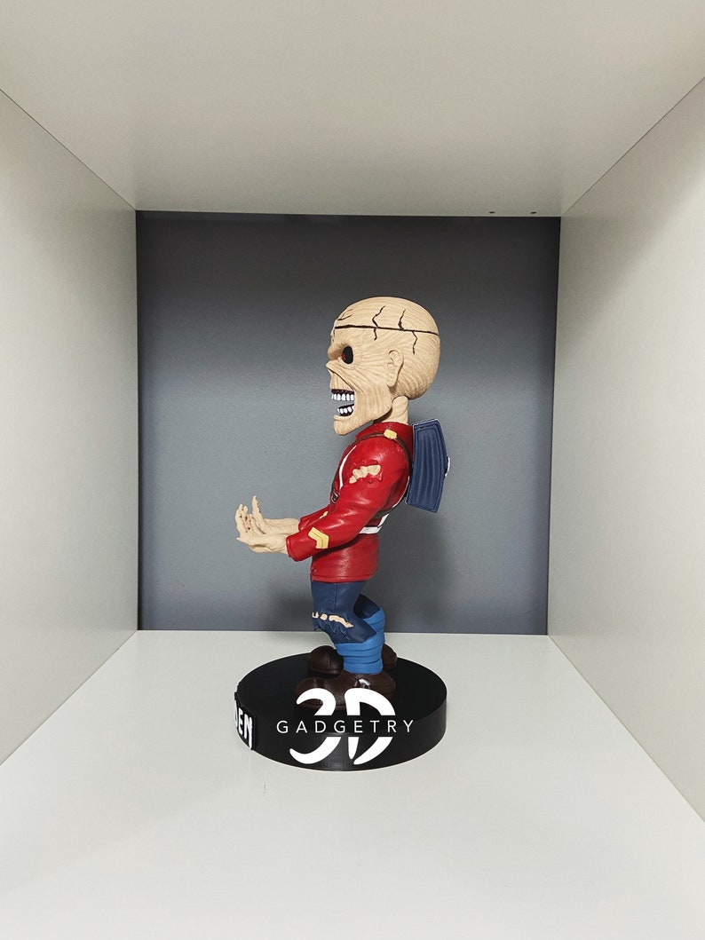 K&ouml;nnte beinhalten: Eine rot-wei&szlig;e Bobblehead-Figur eines Skeletts, das eine rote Jacke, blaue Jeans und einen blauen Rucksack tr&auml;gt. Die Figur steht auf einem schwarzen Sockel mit dem Text "3D GADGETRY".