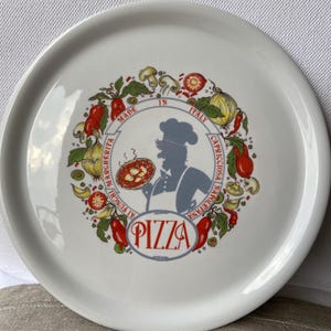 Può includere: Piatto da portata per pizza in ceramica bianca con un'illustrazione colorata. Il design presenta uno chef che tiene in mano una pizza, circondato da verdure e le parole "Made in Italy" e "Pizza".