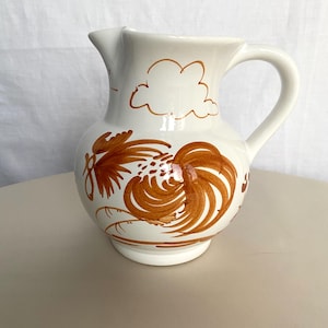 Può includere: Brocca in ceramica bianca con manico e beccuccio curvi. La brocca è decorata con disegni di galli e nuvole marroni. Un oggetto decorativo per la cucina o la sala da pranzo.