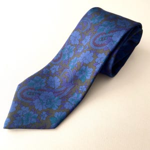Op de afbeelding: Een blauwe stropdas met een bloemenpaisleypatroon. De stropdas heeft een donkerblauwe achtergrond met blauwe, turquoise en paarse bloemmotieven. De stof is glad, mogelijk zijde.