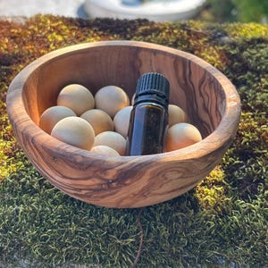May include: A round, natural-grained wooden bowl filled with several smooth, light-coloured wooden spheres and a single dark amber glass dropper bottle with a black cap, positioned centrally within the bowl among the wooden balls.