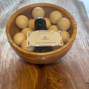 May include: A wooden bowl containing light-coloured wooden spheres and a black bottle. The label on the bottle reads "Oil infused set" and "Vanilla fragrance oil". The bowl is wrapped in clear plastic and rests on a wooden surface.