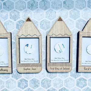 May include: Four wooden pencil-shaped picture frames with adhesive backing. The frames are decorated with a zig-zag pattern and have a space for a photo and a name or phrase. The names on the frames are Oliver Anthony, Sophia Jane, First Day of School, and First Grade.
