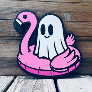May include: A whimsical wooden cutout featuring a friendly ghost floating on a pink flamingo pool float. The ghost is white with black eyes and a smiling mouth. The flamingo and ghost are outlined in black, creating a playful, summery vibe.