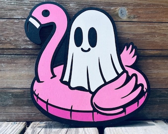 May include: A whimsical wooden cutout featuring a friendly ghost floating on a pink flamingo pool float. The ghost is white with black eyes and a smiling mouth. The flamingo and ghost are outlined in black, creating a playful, summery vibe.