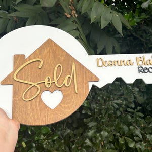 May include: A wooden key-shaped sign with a white circular top and a key-shaped handle. The sign features a house silhouette with the word "Sold" in gold script, a heart cutout, and the name "Deonna Blalock, Realtor".