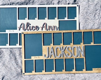 Può includere: Due cornici per foto in legno con sfondo blu e numeri bianchi. La cornice superiore ha il nome "Alice Ann" e la cornice inferiore ha il nome "JACKSON". Entrambe le cornici hanno le parole "Pre K" e "Graduation".