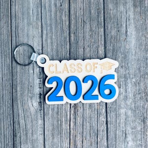 May include: Wooden keychain with the text "CLASS OF 2026" in a blue and natural wood color scheme. The keychain includes a graduation cap graphic and a silver key ring. The design is set against a wooden background.