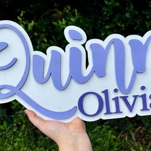 May include: A white wooden sign with the names "Quinn" and "Olivia" written in a purple cursive font.