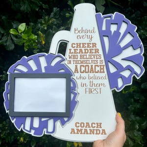 May include: White wooden megaphone with a purple pom-pom on each side. The megaphone has a photo frame and the text "Behind every cheer leader who believes in themselves is a coach who believed in them first. Coach Amanda".