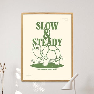 Op de afbeelding: Een ingelijste poster met een cartoon schildpad illustratie. De poster toont de woorden "SLOW & STEADY" in het groen, met de zin "SLOW IS SMOOTH, SMOOTH IS FAST" eronder. De poster staat op een houten bureau met een houten stoel.