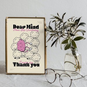 May include: A framed print with the text "Dear Mind, Please Stop Overthinking, I need to sleep, Thank you." The artwork features a pink brain among cartoon sheep. The frame is gold, and a small vase with greenery and glasses are in the foreground.