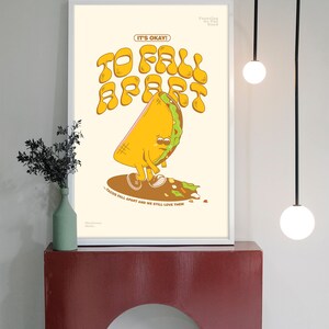 May include: A framed art print featuring a cartoon taco with a sad expression, with the words "TO FALL APART" in large, retro-style lettering. The text "IT'S OKAY!" is above the taco, and "- TACOS FALL APART AND WE STILL LOVE THEM" is below.