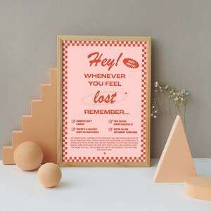 May include: A framed print with a pink background and red checkered border. The text reads "Hey! Whenever you feel lost, remember..." with motivational bullet points. Decorative geometric shapes and spheres are in the foreground.