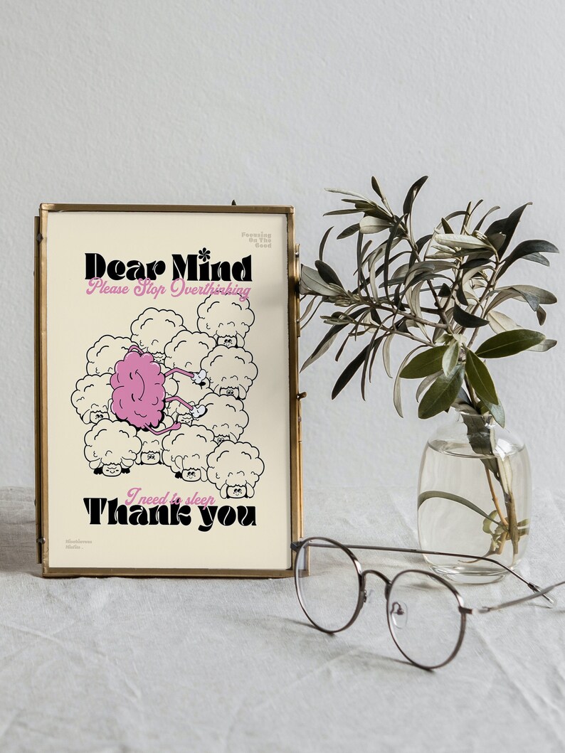 May include: A framed print with the text "Dear Mind, Please Stop Overthinking, I need to sleep, Thank you." The artwork features a pink brain among cartoon sheep. The frame is gold, and a small vase with greenery and glasses are in the foreground.