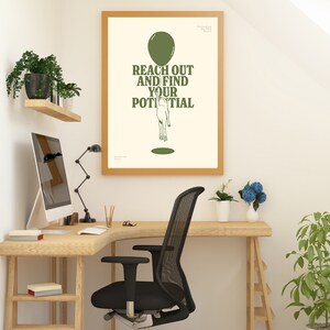 Pu&ograve; includere: Un poster incorniciato con la scritta "REACH OUT AND FIND YOUR POTENTIAL" in verde, sopra un'illustrazione di una persona che raggiunge un palloncino. Il poster &egrave; in una cornice di legno chiaro, esposto in un ambiente di home office con una scrivania in legno e una sedia da ufficio nera.