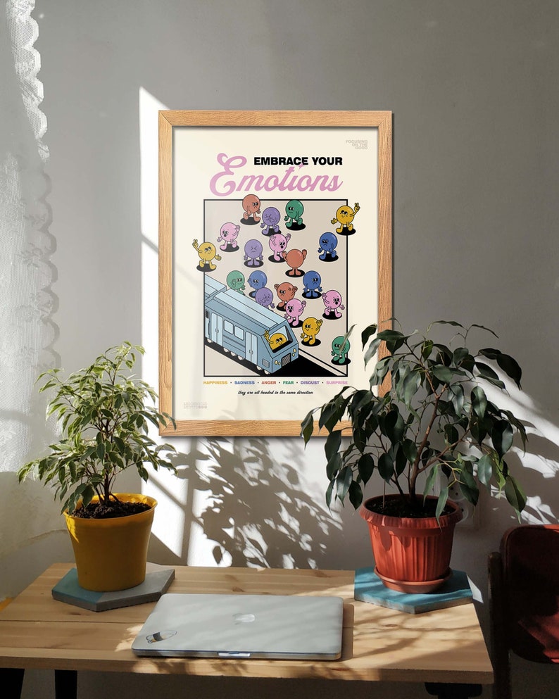 May include: A framed poster with the text "EMBRACE YOUR EMOTIONS" and cartoon figures representing different emotions. The poster is in a light wood frame and is displayed on a wooden desk with plants and a laptop.