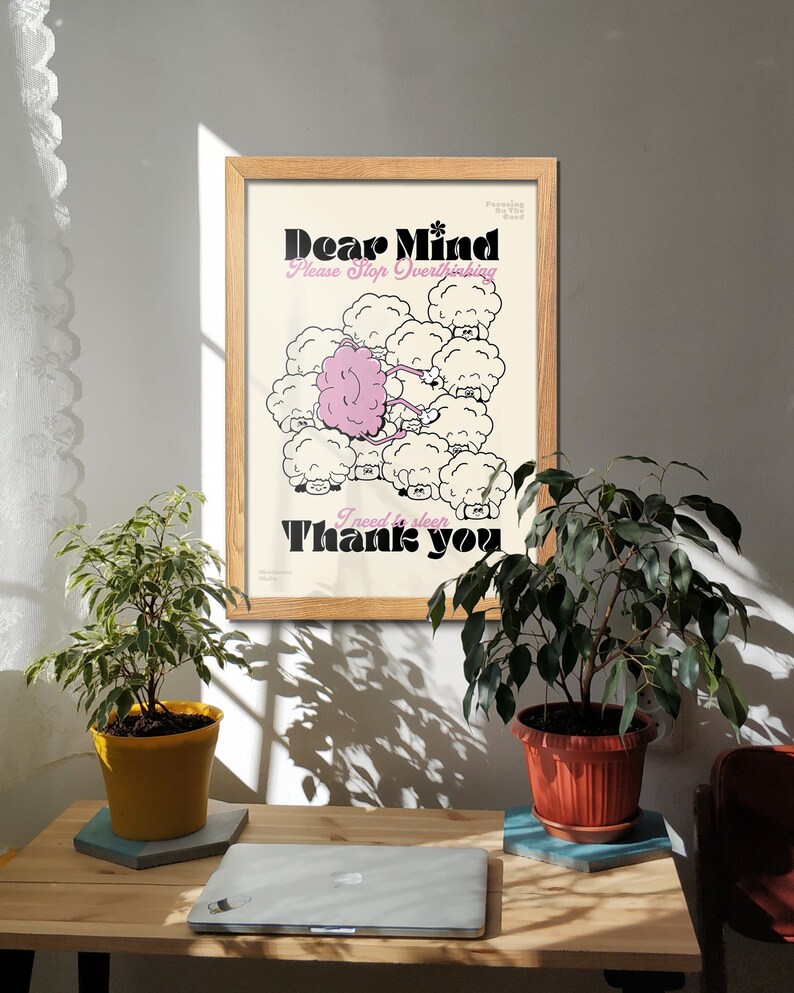 May include: A framed poster with the text "Dear Mind Please Stop Overthinking Thank you" in black and pink. The artwork features a pink brain surrounded by white sheep-like figures. Two potted plants and a laptop are on a wooden desk.