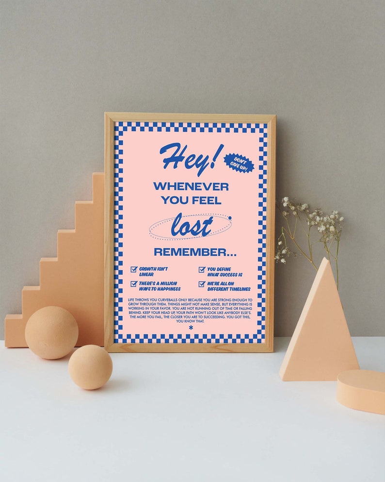 May include: A framed poster with a pink background and blue text that reads "Hey! Whenever you feel lost, remember..." The poster includes a blue checkered border and a small "Don't Give Up!" sticker. Decorative geometric shapes are in the foreground.