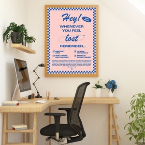 May include: A framed poster with a pink background and blue checkered border. The poster reads "Hey! Whenever you feel lost, remember..." with a list of inspirational reminders. The poster is displayed on a wooden desk with a black office chair.