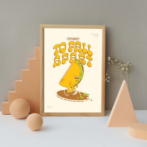 May include: A framed print featuring a cartoon taco character with the text "TO FALL APART" and "IT'S OKAY!". The taco is yellow with a green border, brown shoes, and a sad expression. The print is in a wooden frame.