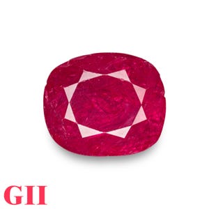 May include: A cushion-cut ruby gemstone with a vibrant red hue. The gem features a star-like pattern in the center, with facets that reflect light. The letters "GII" are displayed in red at the bottom.
