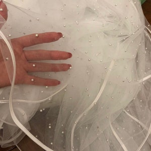 Puede incluir: Velo de tul blanco con una capa de cinta de raso blanco y adornos de perlas dispersas.