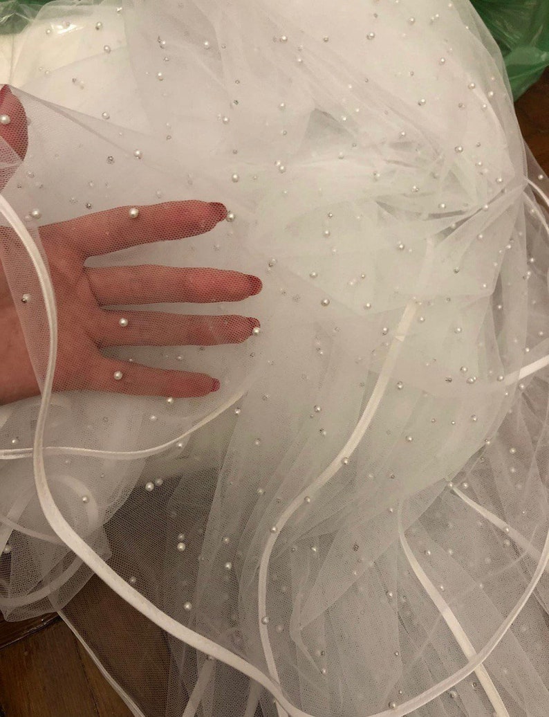 Puede incluir: Velo de tul blanco con una capa de cinta de raso blanco y adornos de perlas dispersas.