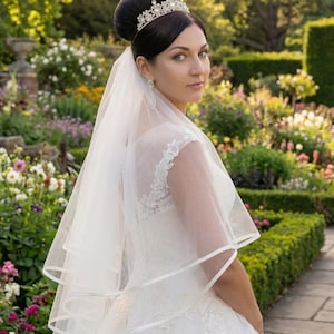 May include: A white wedding dress featuring intricate lace detailing on the bodice and a layered tulle skirt. A sheer, multi-tiered veil with a satin ribbon edge is attached to the hair, which is styled in an updo with a silver crystal tiara.