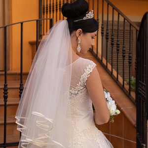 Puede incluir: Una novia con un vestido de novia blanco con detalles de encaje y un velo. Lleva un ramo de flores blancas y una tiara en la cabeza.