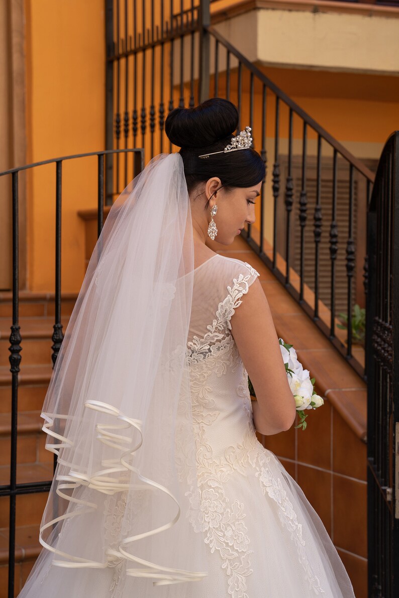 Puede incluir: Una novia con un vestido de novia blanco con detalles de encaje y un velo. Lleva un ramo de flores blancas y una tiara en la cabeza.