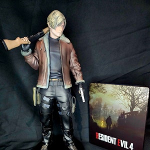 May include: A collectible figurine of Leon S. Kennedy from Resident Evil 4, holding a rifle and a pistol. The figurine is standing on a black base with the words "Leon S. Kennedy" printed on it. The figurine is next to a video game case for Resident Evil 4.